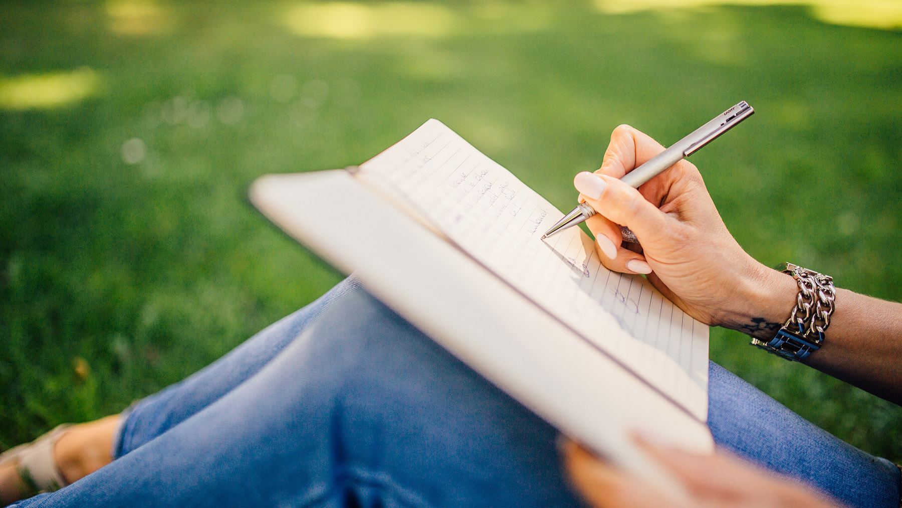 Stock photo of someone writing in a journal type book while sitting outdoors on a sunny day (Image by RawPixel.com)
